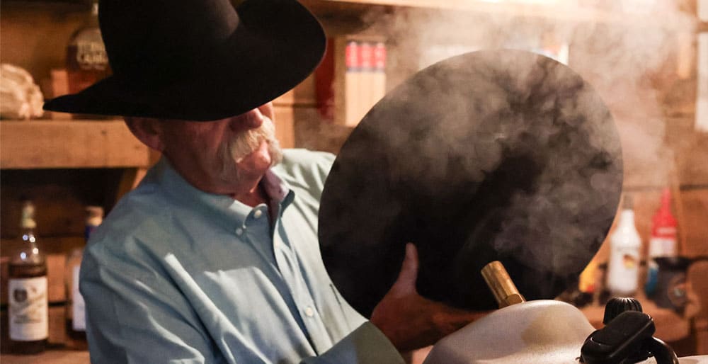 Un vaquero con un sombrero negro, sosteniendo un sombrero negro frente a una máquina de vapor, calentando el sombrero con vapor.